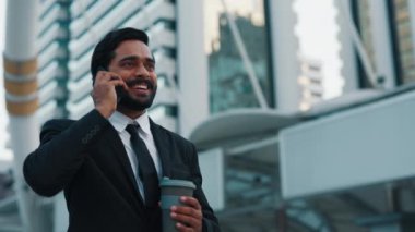 Mutlu iş adamı, elinde sürdürülebilir içki bardağı tutarken içten bir şekilde gülerek telefon konuşmasına daldı. Yakışıklı erkek ofis merkezinin yanında duruyor ve hoş sohbetin tadını çıkarıyor..