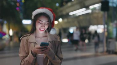 Kadın turist şehrin etrafında yürüyor ve Noel Baba şapkası takarak yılbaşı arifesinde telefondan kayıyor. Kış Tatillerinde Sosyal Ağları Çalan Güzel Pozitif Kadın Telefonu Kullanıyor