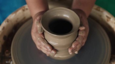 Çamurlu bir nesneyi palmiyelerle tutarken dönen makinenin içindeki nesneyi yapan karışık ırktan birinin kirli ellerinde üst görünüm. Bilinmeyen insan çömlek tekeriyle çalışıyor ve sürahi şeklinde.