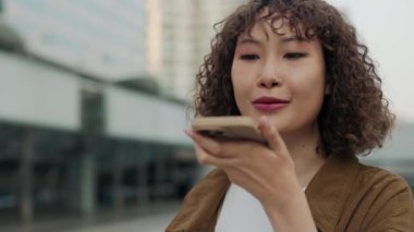 Merkezde dikilirken telefonuna sesli mesaj kaydeden kıvırcık saçlı mutlu bir Asyalı kadın. Burnu piercingli genç bir kadın arka plandan hoparlöre konuşuyor. Halk ve Teknoloji Konsepti