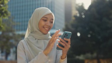 Gülümseyen Çekici Kadın dışarıda duruyor ve tesettürlü cep telefonuyla uygulamaları kaydırıyor. Geleneksel Giysilerdeki Şirin Bayan Gülümsüyor, Açık havada Akıllı Telefon 'da Haberleri Çalıyor. Halk ve Teknoloji