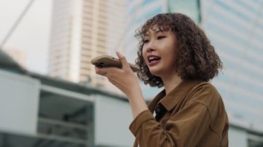 Kıvırcık saçlı mutlu Asyalı kız dışarıda dikilirken telefonuna sesli mesaj gönderiyor. Ai 'yi veren genç Asyalı kadın şehrin arka planıyla telefonda konuşuyor. Halk ve Teknoloji Konsepti