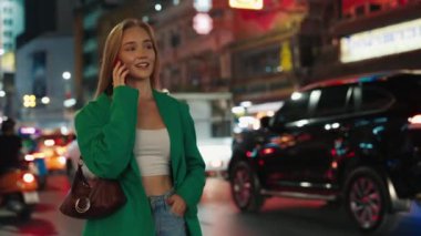 Günlük kıyafetlerle gezen mutlu sarışın kadın portresi. Şehir caddesinde akıllı telefondan konuşuyor. Yeşil ceketli bir kadın akşamları cep telefonuyla gezintiye çıkar.