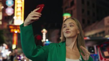 Gece caddesinde cep telefonuyla selfie çeken çekici sarışın kadın portresi. Genç bayan, akşam kenti bölgesinde fotoğraf çekmek için saç ayarlıyor.