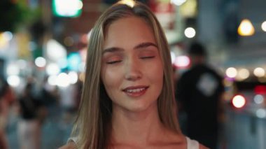 Aydınlanmış gece caddesinde kameraya gülümseyen güzel bir genç kadının portresi. Uzun sarı saçlı mutlu kadın, şehir merkezinde olumlu duygular ifade ediyor.