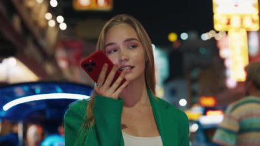 Güzel sarışın bir kadının portresi akıllı telefondan sesli mesaj yolluyor ve neon ışıklı gece şehrinin ortasında duruyor. Kadın, modern akşam megapolis 'inde cep telefonuyla iletişim kurar.