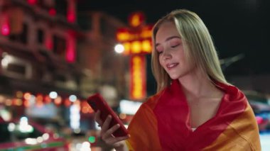 Meşgul bir gece caddesinde akıllı telefon kullanan gökkuşağı bayrağıyla güzel bir kadının portresi. Gülümseyen sarışın kadın cep telefonuyla LGBT topluluk desteği mesajı gönderiyor