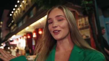 Gece şehir sokağındaki kameraya bakan takipçilere el sallayan mutlu kadın portresi. Şehir merkezindeki modern alışveriş merkezinin olumlu kadın blog kayıtları incelemesi. POV