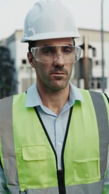Güvenlik yeleği giymiş insan mühendisinin portresi kameraya bakıyor. Koruyucu göz ve kask giyen erkek müteahhit proje alanında verimli bir güne hazırlanırken kendine güven yayıyor. Dikey çekim