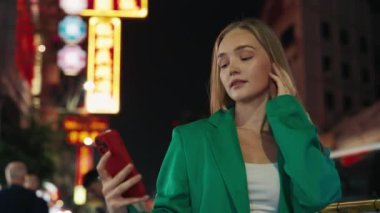 Kendine güvenen bir kadının portresi cep telefonuyla modern şehir geçmişiyle selfie çekiyor. Canlı spor ceket giyen güzel sarışın, şehirde sosyal medya için görsel içerik sağlıyor.