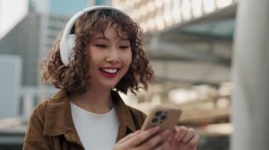Güzel Pozitif Kız, elinde kulaklıkla telefonda sohbet ederken sokakta yürüyor. Arka planda sosyal medyada gezinen neşeli ve neşeli bir kadın. Halk ve Teknoloji