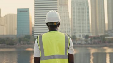 Kasklı ve sarı yelekli Afro-Amerikan müteahhit şehrin arka manzarasında. İnsan müteahhiti nehir kıyısındaki yüksek binalara bakıyor. Kentsel altyapı geliştirme