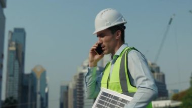 Akıllı telefondan konuşan meşgul bir mühendisin portresi şehirdeki yüksek binalarda yürüyor. Sert şapkalı erkek profesyonel, güneş paneli sorunlarını telefonda çözüyor