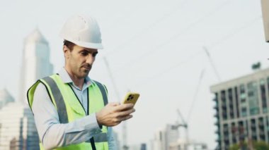Sert şapkalı bir mühendisin portresi, akıllı telefona bakarken muzaffer jestler yapıyor, elektrik hatlarının yanında duruyor. Güvenlik yeleği giyen erkek işçi, başarı onayını kutluyor