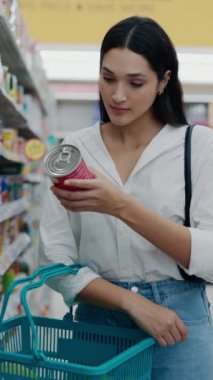 Dikey video. Dükkanda malzemeleri okumak için paketlenmiş bir ürünü elinde tutan hoş bir kadın. Süpermarketteki güzel bayan yiyecekleri topluyor. Alışveriş Konsepti