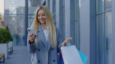 Alışveriş yaptıktan sonra iş kadını, paketlerle gidiyor ve akıllı telefonundaki sosyal ağlar aracılığıyla kayıyor. Paketleri tutarken akıllı telefon kullanan genç bayan. Reklam ve Teknoloji Konsepti