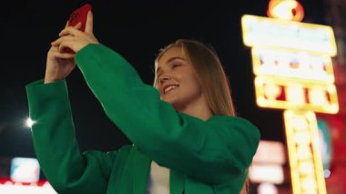 Güzel bir kadının portresi akıllı telefondan fotoğraf çekiyor. Gece şehir caddesinde duruyor. Pozitif kadın blogcu, şehir merkezinde akşamları cep telefonuyla eğleniyor.