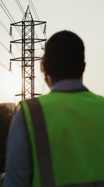 İnsan mühendisin arka görüntüsü, elektrik hatlarının yanında duran kaskı takıyor. Yansıtıcı yelek giyen erkek müfettiş, potansiyel olarak tehlikeli ortamlarda işyeri güvenliği uygulamalarının önemini vurguladı