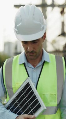 Güç fabrikasının yanında duran küçük güneş paneli tutan insan mühendisinin portresi. Güvenlik kasklı erkek müteahhit kameraya bakarak sorumlu enerji tüketimini teşvik ediyor