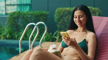 Bikinili gülümseyen kadın, şezlongda uzanan akıllı telefonla mesajlar yolluyor. Havuz kenarında hindistan cevizi içkisi içen güzel bir kadın yatıyor. Bayan yüzme havuzu yakınında tatilden hoşlanır.