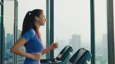 Genç sporcu kadın spor salonundaki panoramik pencerenin yanındaki koşu bandında koşuyor. Mutlu kadın sağlıklı yaşam tarzını geliştirmek için kardiyo çalışması yapıyor. Eşofmanlı zayıf kadın atletik enerji gösteriyor. Yakınlaştır