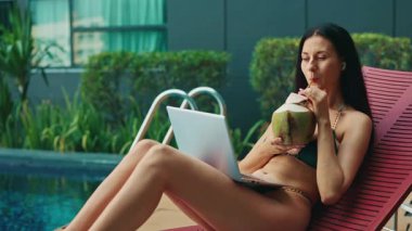 Genç bir kadın yüzme havuzu yakınında hindistan cevizi kokteyli içerek dizüstü bilgisayar kullanıyor. Kadın, tropik tatil beldesinde verimlilik ve gevşemeyi dengeler. Bayan güvertede dinleniyor ve sohbet ediyor.