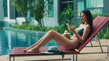 Kırmızı şezlonga uzanmış akıllı telefondaki bikinili çekici esmer kadın. Güzel bayan turist havuz kenarında güneşleniyor. Bayan egzotik otel yüzme havuzunun yanında dinleniyor.