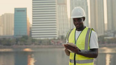 Şehrin rıhtımında elinde tablet tutan mutlu Afrikalı Amerikalı müteahhit portresi. Kasklı erkek mühendis şehir bölgesindeki inşaat verilerini kontrol ediyor. Mimari Geliştirme