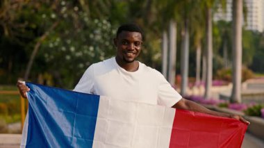 Büyük bir Fransız bayrağı taşıyan Afro-Amerikan bir adam parkta dikilirken kameraya bakıyor. Günlük tişörtlü adam Fransa 'da yaya bölgesinde düzenlenen tarihi etkinliği onurlandırdı.