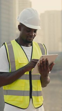 Kasketli işçilerin portresi şehir seti üzerinde duran akıllı tablet üzerindeki çalışma programını inceliyor. Yansıtıcı yelek giyen Afrikalı Amerikalı erkek müteahhit materyalleri internette tarıyor