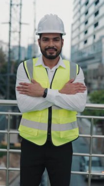 İnşaat alanında kask ve çelik yelek giyen kendinden emin mühendisin portresi. Gülümseyen müteahhit köprüde tamamlanmamış binaya karşı kollarını kavuşturur. Dikey çekim