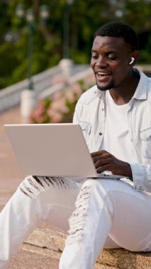 Kulaklıklı Afro-Amerikalı adam laptopa bakarken video aracılığıyla konuşuyor. Günlük kıyafetli neşeli adam parktaki merdivenlerde oturarak online konferansa katılıyor. Dikey çekim