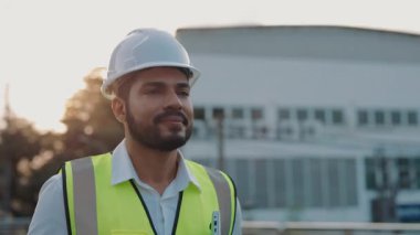 Günbatımında binaların önünde dizüstü bilgisayarla yürüyen olgun bir inşaatçının portresi. Güvenlik yeleği giyen erkek mühendis akşam iş yerinden ayrılıyor. İş mesleği