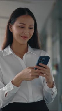 Dikey video. Sosyal medyada cep telefonu taraması yapan çekici, pozitif iş kadını. Hoş bir kadın içeride mesajlaşıyor, cep telefonunda kayıt uygulamaları var. Halk ve Teknoloji Konsepti