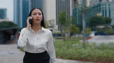 Beyaz bluzlu bir kadın ciddi bir ifadeyle akıllı telefonuyla konuşuyor. Şehir merkezinde gökdelenlerin zeminine karşı yürüyor, iş iletişimi kuruyor..