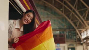 Yolcu arabasının camından sarkan gökkuşağı bayrağı tutan genç Asyalı transkadın portresi. LGBT gururunu gösteren mutlu bayan turist trenle seyahat ediyor. Boşluğu kopyala