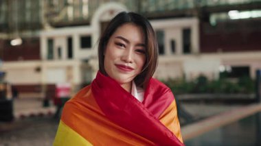 Tren istasyonunda parlak gökkuşağı bayrağıyla sarılmış güzel Asyalı lezbiyenin portresi. Olumlu eşcinsel kadın turist, LGBTQI toplum eşitliğini tren taşımacılığıyla destekliyor
