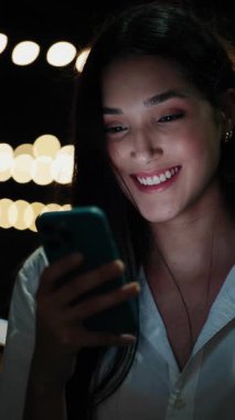 Dikey video. Genç kız ayakta duruyor ve gülümsüyor, dışarıdaki telefonuna bakıyor. Güzel, Olumlu Genç İş Sahibi Akıllı Telefonunu Kullanıyor. Gece vakti. Halk ve Teknoloji Konsepti