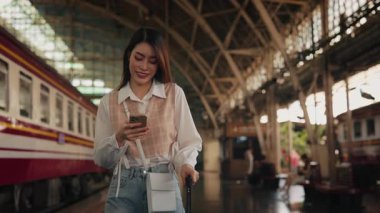 Demiryolu platformunda bavul taşıyan akıllı telefondan mesaj atan neşeli Asyalı transseksüel portresi. Tren istasyonunda uçağa binmeden önce telefonla gülümseyen lezbiyen mesajları