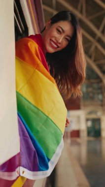 Demiryolu arabasının camından sarkan dişlek gülüşlü ve gökkuşağı bayrağıyla Asyalı transkadın portresi. Eşcinsel kadın gezgin LGBT gururlu yolculuk trenini paylaşıyor. Dikey çekim