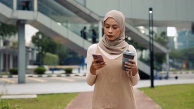 Malezyalı tesettürlü iş kadını portresi açık havada akıllı telefon ve kağıt bardak tutuyor. Genç Müslüman kadın, şehir parkında cep telefonuyla internette gezinmeyi seviyor