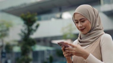 Akıllı telefon üzerinde tesettürlü metinleri olan gülümseyen Malezyalı genç bir kadının portresi. Mutlu muslin dişisi şehirde online iletişim için mobil teknolojiye sahip.