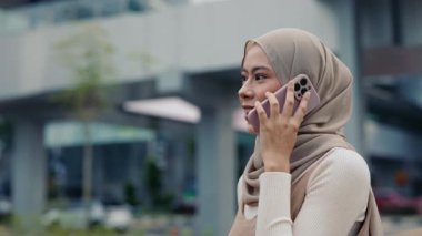 Tesettürlü neşeli genç kadın portresi şehir sokaklarında telefon konuşmalarından hoşlanıyor. Malezyalı kadın şehir ortamında bağlantı ve kültürel ifade sergiliyor