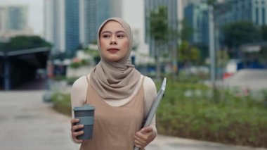 Başörtülü Müslüman kadın portresi şehir manzarasına karşı dizüstü bilgisayar ve yeniden kullanılabilir kahve fincanı taşıyor. Genç Malezyalı kadın çalışanlar şehirde verimli bir iş gününe hazır.