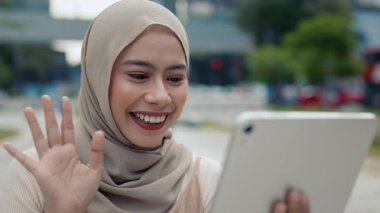 Türbanlı mutlu genç Malezyalı kadının yakın portresi sokaktaki dijital tablet standlarında video çağrısı yapıyor. Müslüman kadın ekranda el sallıyor ve şehirde online iletişimin keyfini çıkarıyor