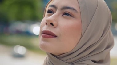 Başörtüsü takan Malezyalı genç bir kadının portresi açık havada düşünceli bir ifadeyle yukarı doğru bakıyor. Müslüman bir kadın şehirde umut ve tefekkür duyguları taşır.