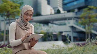Malezyalı tesettürlü kadın portresi, tablet üzerindeki stilus ile birlikte şehir yeşillikleri arasında dikkatlice çiziliyor. Odaklanmış Müslüman kadın, yaz kentinde mobil cihazlara dijital sanat çiziyor
