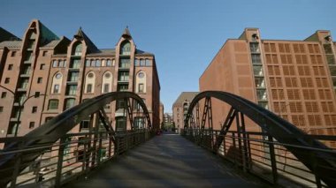 HAMBURG, ALMANY - 23 Eylül 2024: Kanal üzerindeki yaya köprüsü güzel bir günde Speicherstadt ilçesini birbirine bağlar. Şehrin sanayi bloğundaki açık gökyüzünün altındaki tarihi depolarda binalar ve viyadük