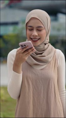 Geleneksel tesettürdeki mutlu Malezyalı kadın portresi şehir sokaklarında dururken sesli mesaj gönderiyor. Genç Müslüman kadın şehrin dikey çekimlerinde cep telefonuyla çevrimiçi sohbet ediyor