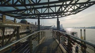HAMBURG, Almanya - 23 Eylül 2024: Krom parmaklıklı ve Elbe Nehri üzerinde şeffaf çerçeveli çatısı olan yaya köprüsü. Günbatımında St. Pauli Landungsbrcken 'in modern viyadük simgesi. Şehir portu